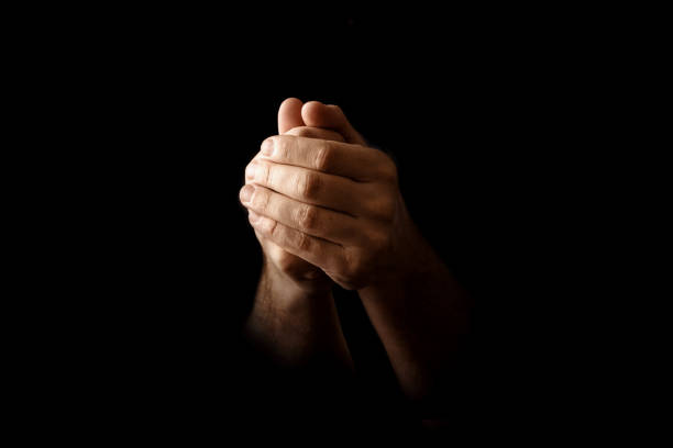 Men's hands in prayer on a black background. The concept of faith, prayer, mourning, forgiveness, confession.
